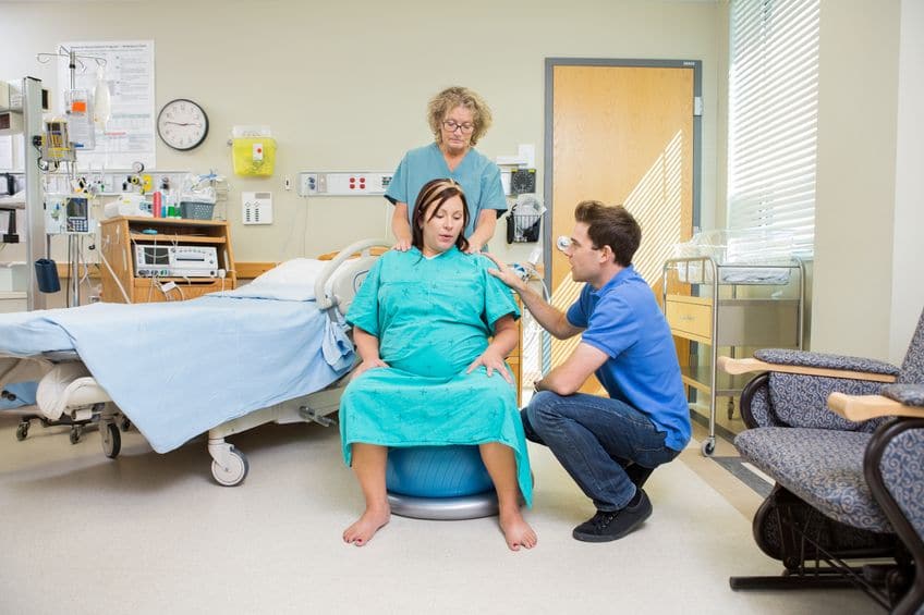A pregnant woman in a delivery room sitting on a birth ball being massage by a Doula. 