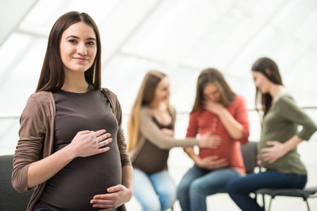  beautiful pregnant woman is looking at the camera. happy pregnant women are talking together at antenatal class at the hospital on background.
